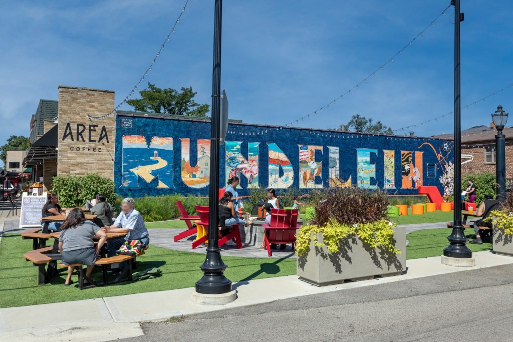 People sitting at picnic tables and red chairs in a vibrant outdoor seating area near Area Coffee, with a colorful mural spelling 'Mundelein' in the background.
