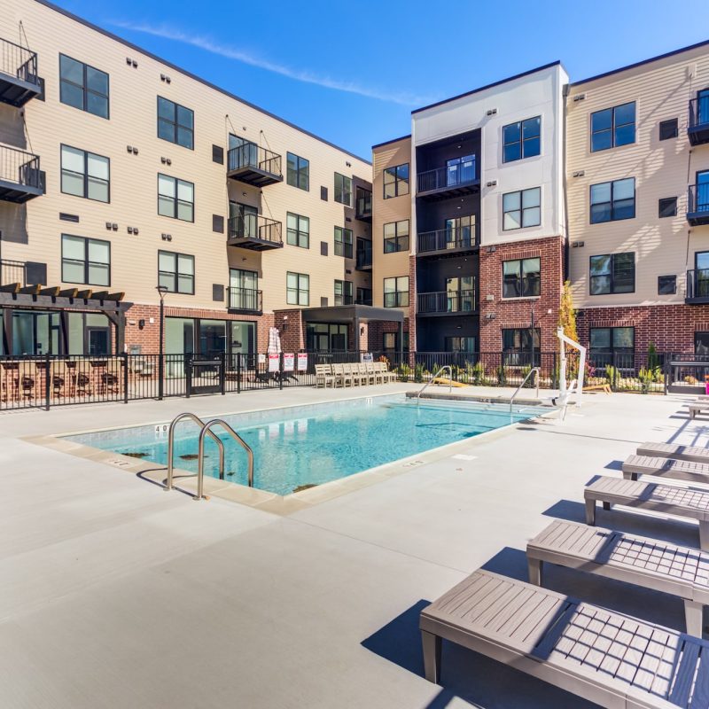 Outdoor swimming pool courtyard with lounge chairs and spacious deck surrounded by four-story Station 250 apartment buildings with balconies under clear blue sky in Mundelein IL